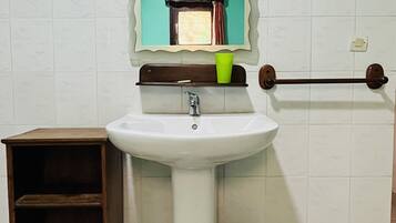 A bathroom with a white sink, a mirror, a shelf with a green cup, a wooden shelf unit, and a black trash bin.