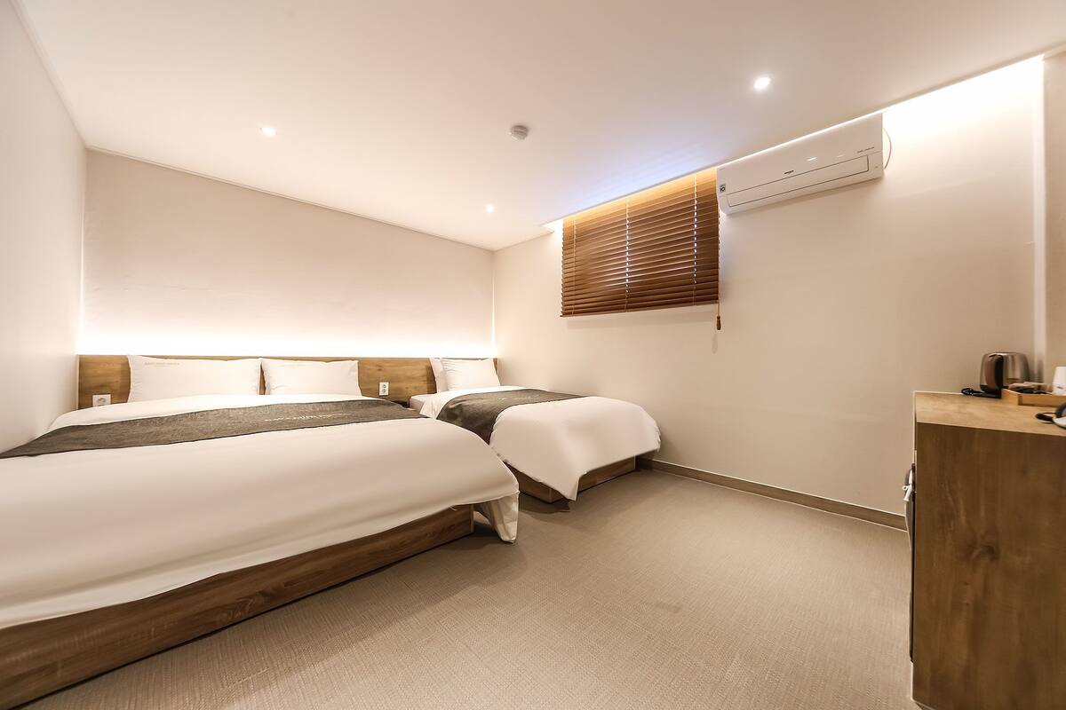 A modern hotel room with a large bed, a smaller bed, a wooden headboard, a bedside table, a desk, and an air conditioning unit.