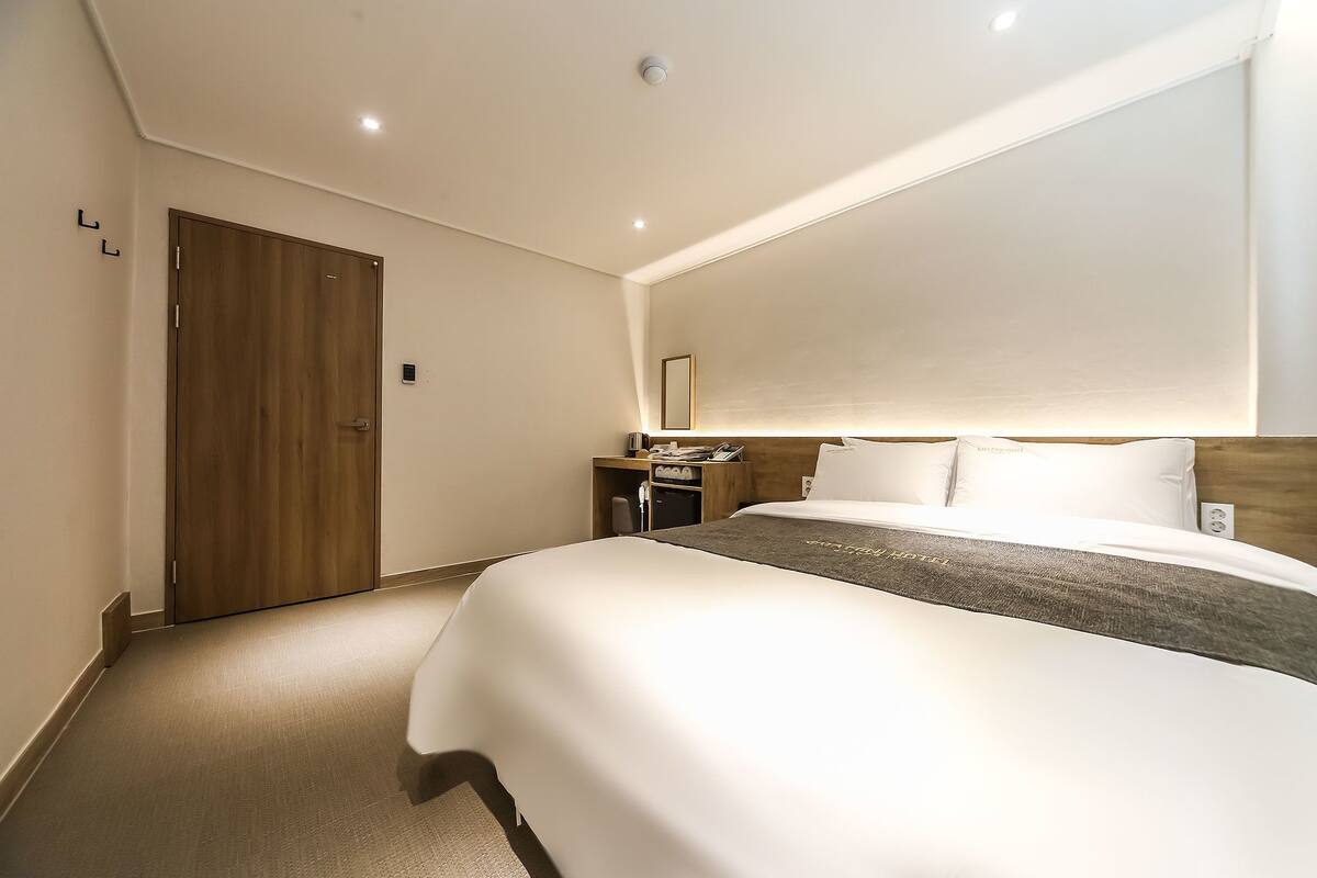 A hotel room with a large bed, a wooden door, a bedside table with a lamp, and a small desk with a chair.