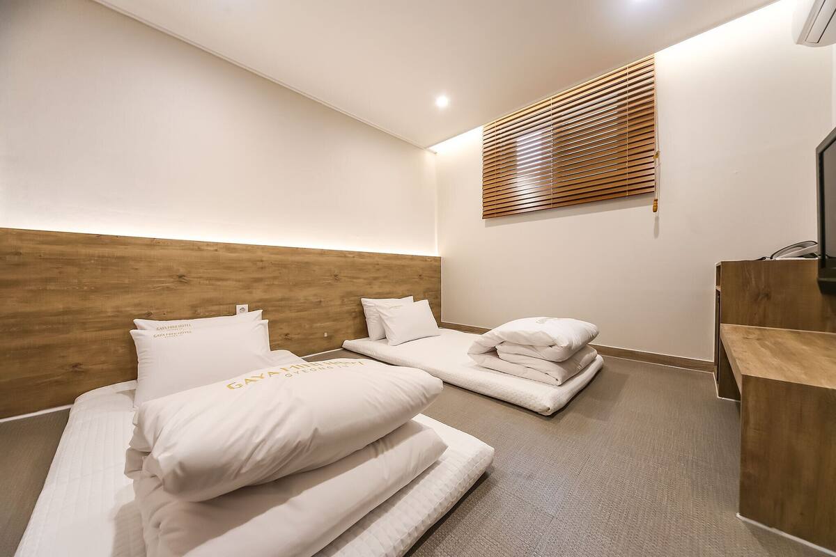 A hotel room with two beds, a wooden headboard, a wooden desk, and a window with blinds.