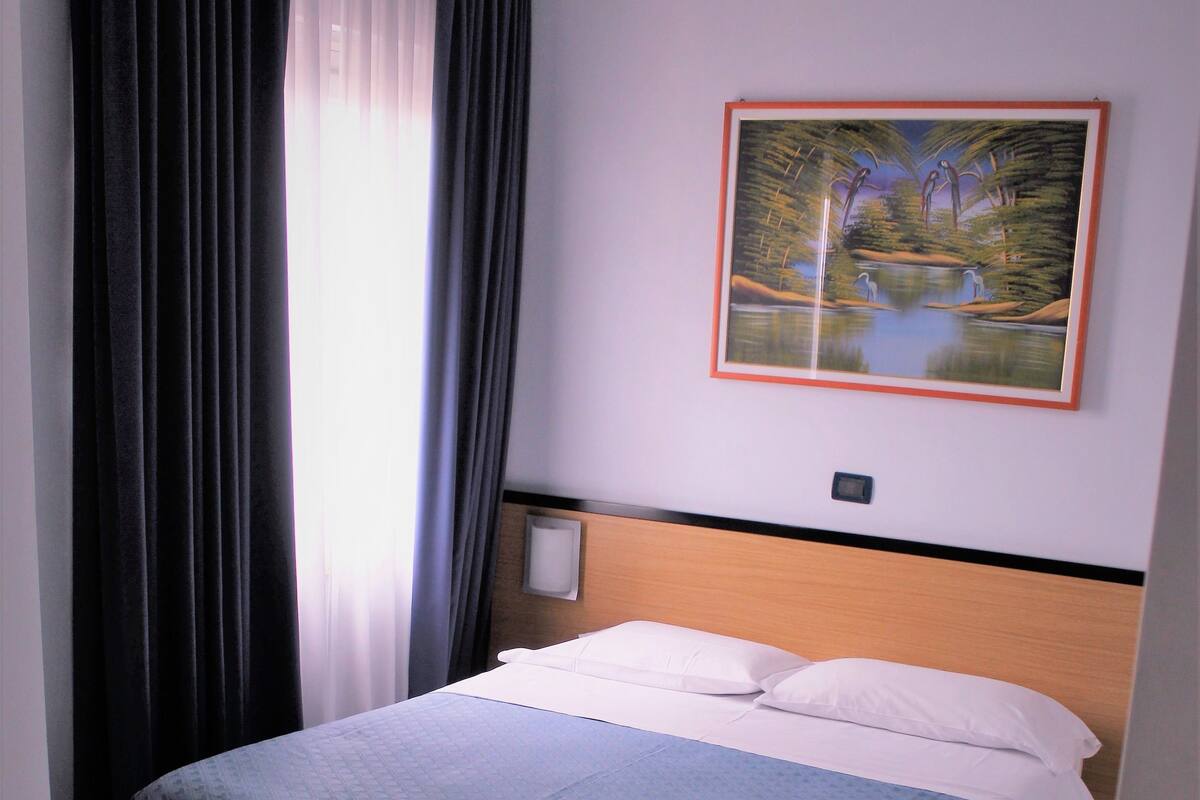 A hotel room with a bed, a painting on the wall, and a window with curtains.