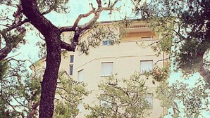 Um edifício de vários andares com fachada bege, rodeado por árvores e um jardim bem cuidado.