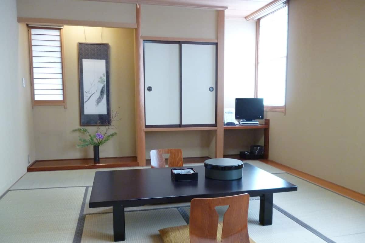 A traditional Japanese room with tatami flooring, a low table, a chair, a television on a shelf, and a framed artwork on the wall.