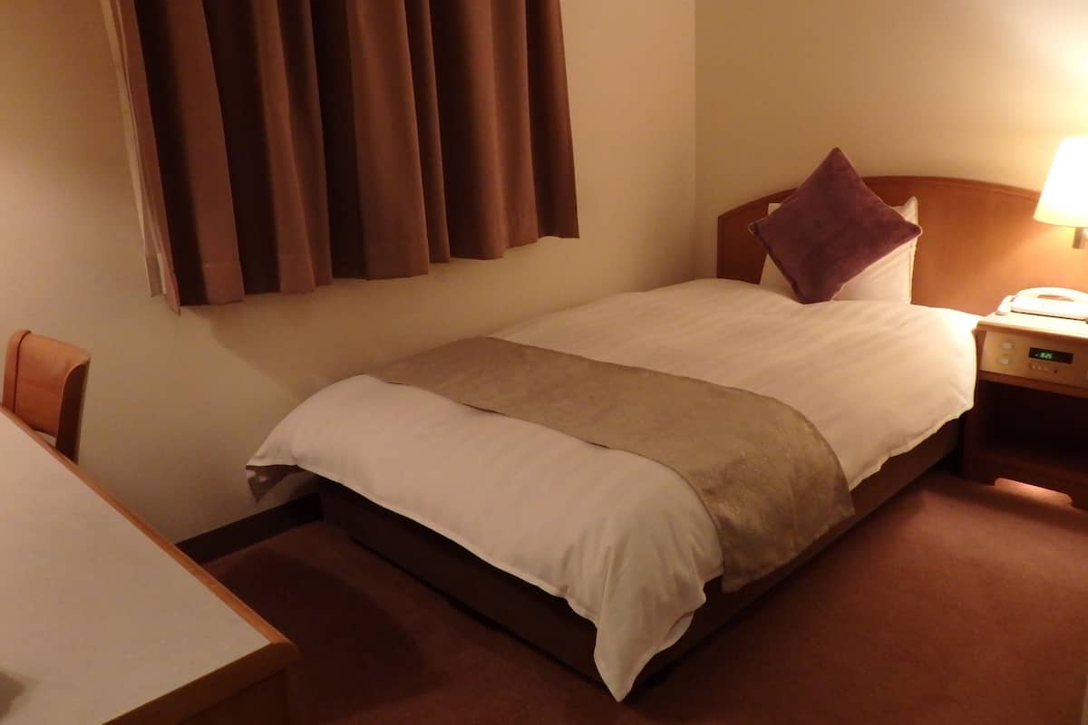 A hotel room with a bed, a nightstand with a phone, a chair, and a desk.