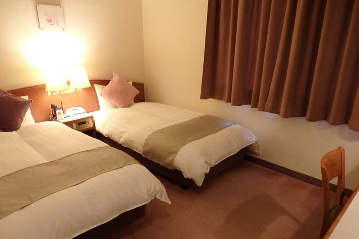 A hotel room with two beds, a nightstand, a lamp, a chair, and a window with curtains.