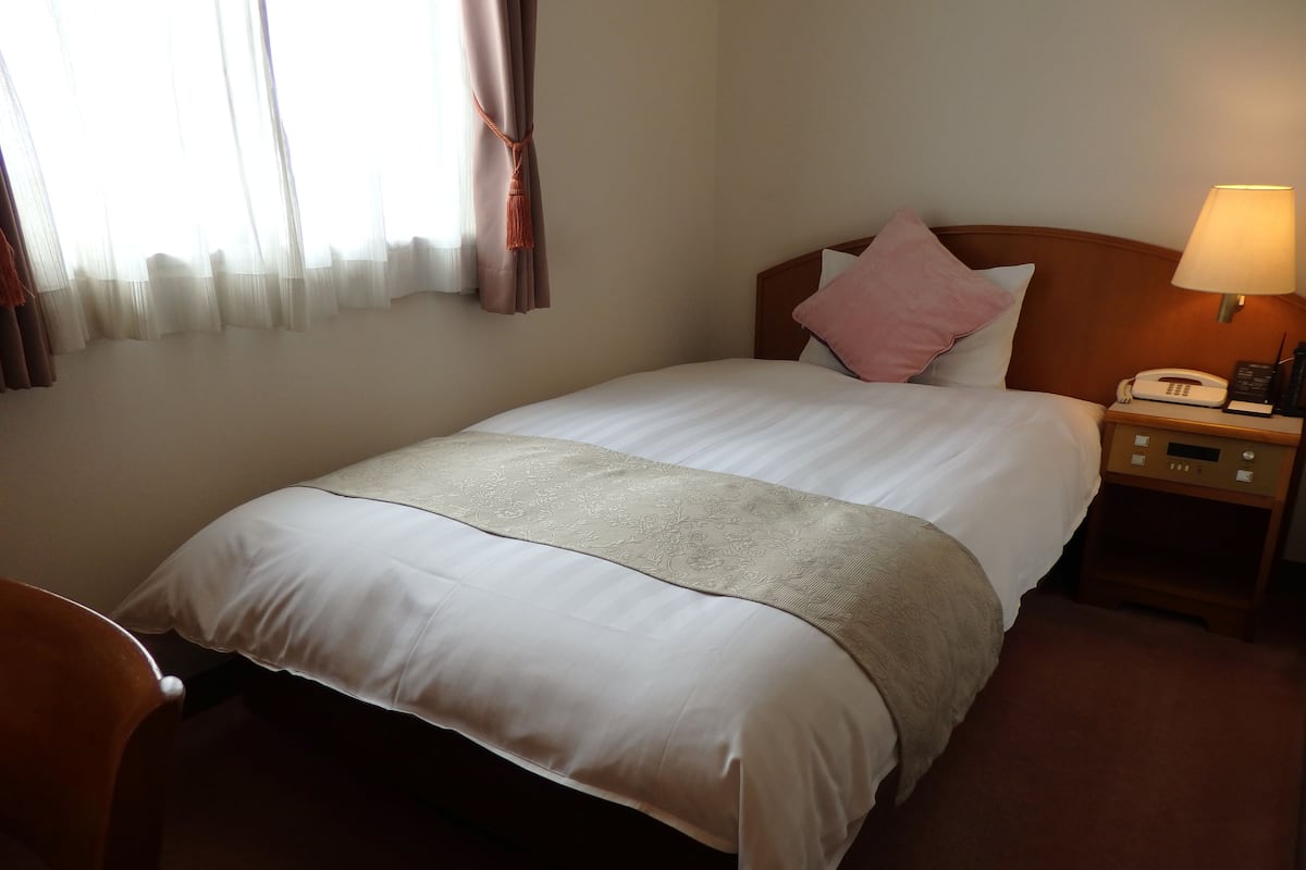 A hotel room with a neatly made bed, a bedside lamp, a nightstand with a phone, and a window with sheer curtains.