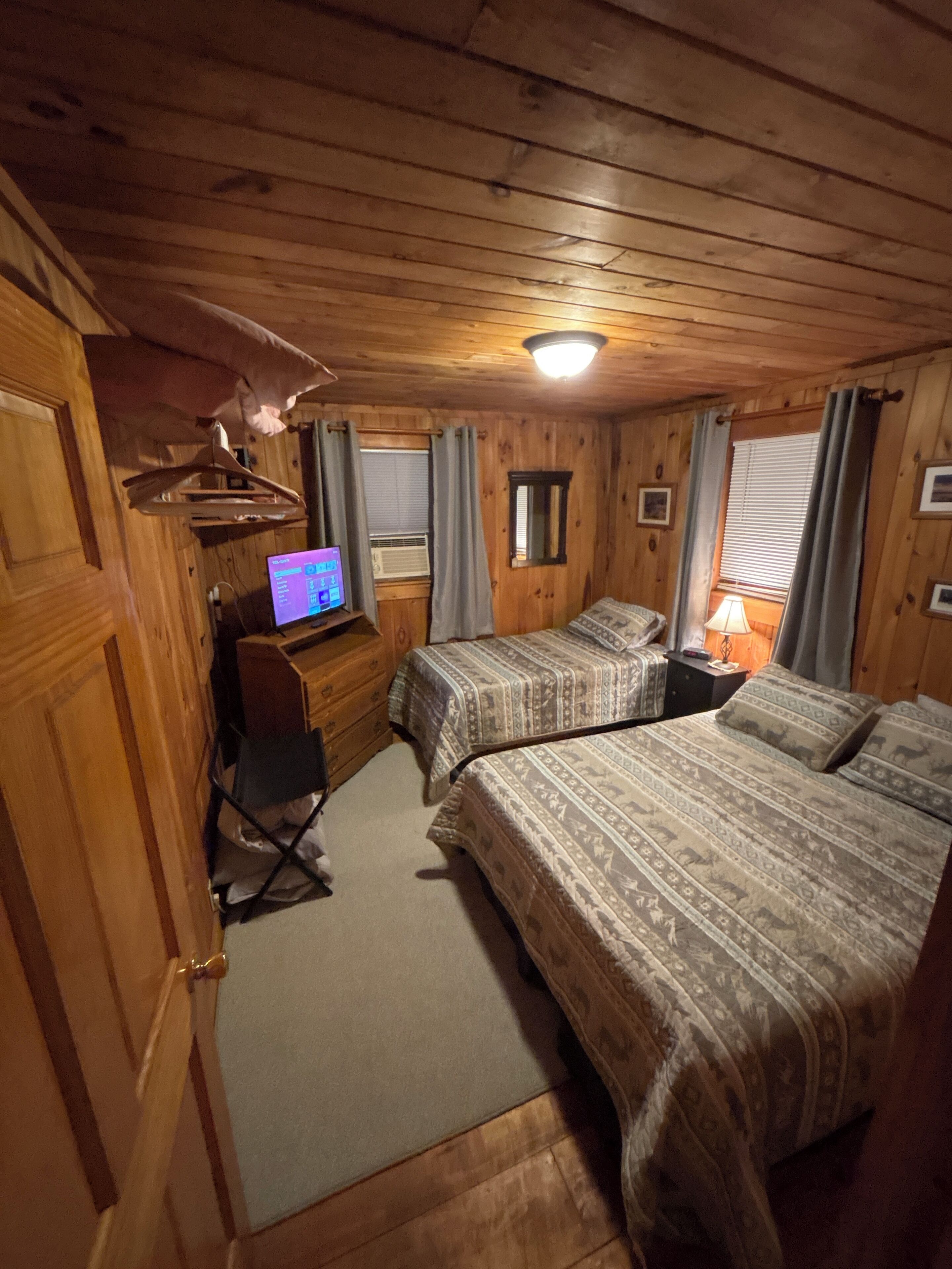 A cozy cabin-style room with two beds, a desk with a computer, a hanging coat rack, and a window with curtains.