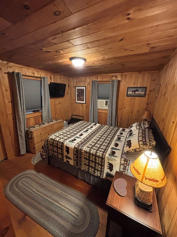 A wooden cabin bedroom with a bed, bedside tables, a lamp, and a window with curtains.
