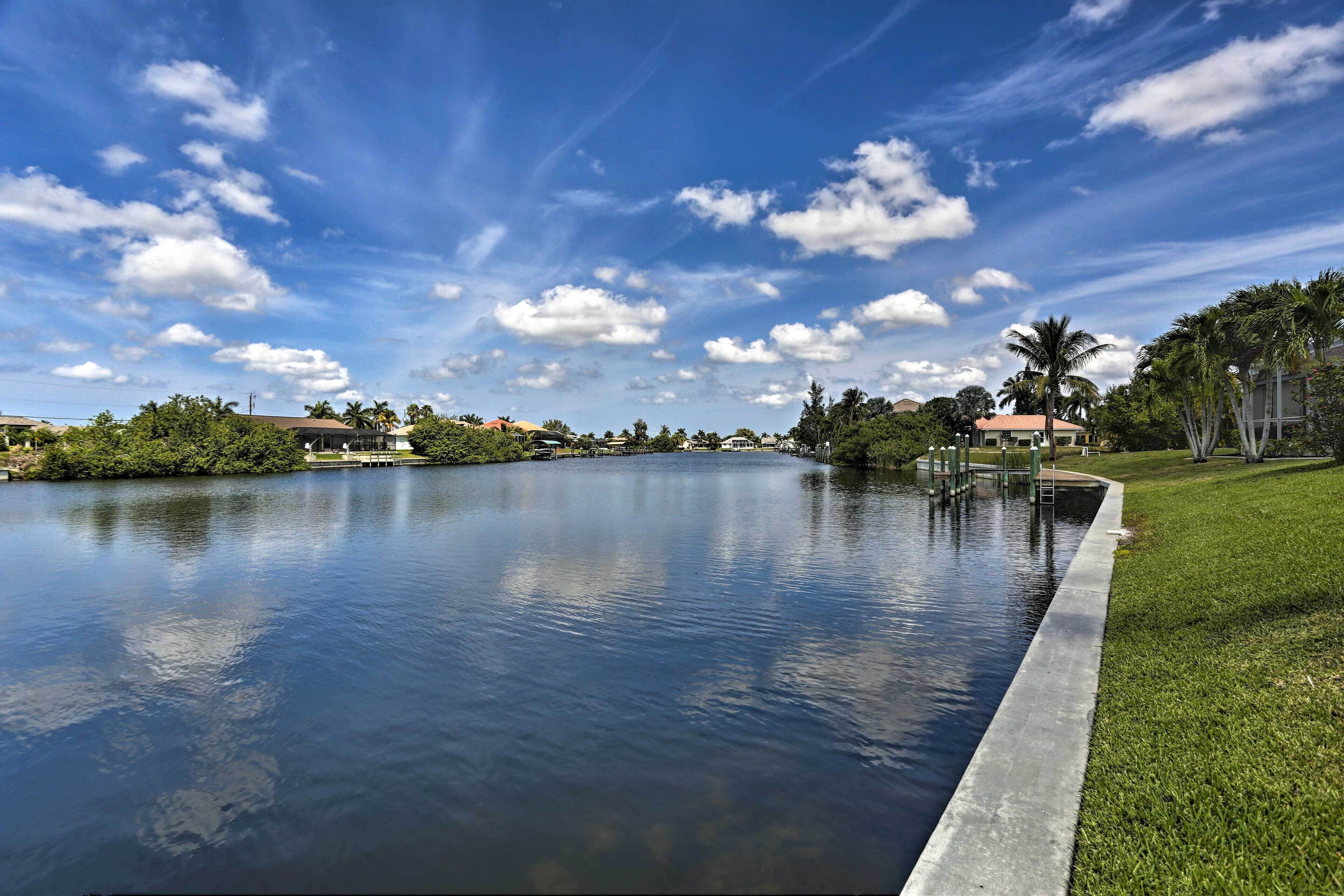 Un lago tranquilo con un paseo marítimo de hormigón a lo largo de la orilla, rodeado de vegetación y edificios al fondo.