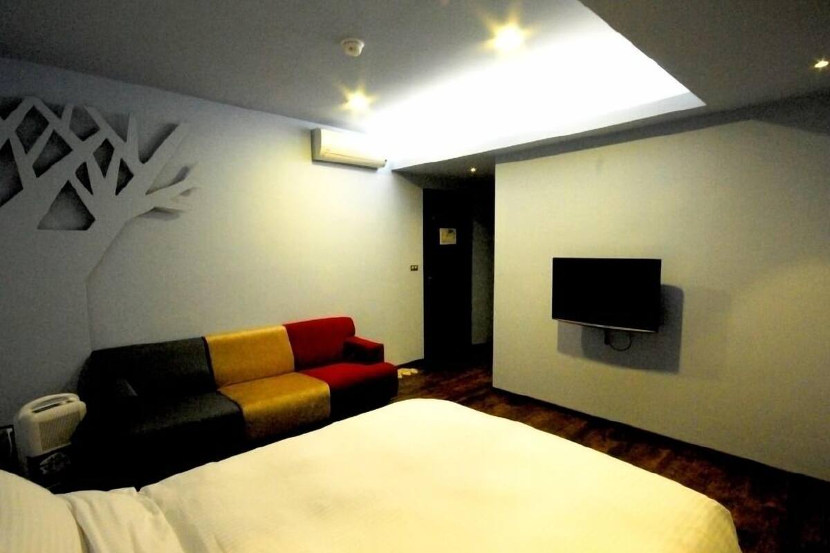 Une chambre d’hôtel avec un lit, un canapé rouge et jaune, un téléviseur à écran plat fixé au mur et un luminaire au plafond.
