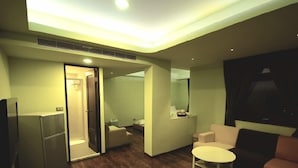 A modern living room with a flat-screen TV, a small round table, a sofa, and a separate bedroom with a bed and a bathroom. - J.F Sleepless Ones Garden Hotel (New Taipei City)