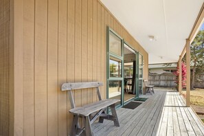 Una terraza de madera con un banco, un ventanal de vidrio y un espacio cubierto con una mesa y sillas.