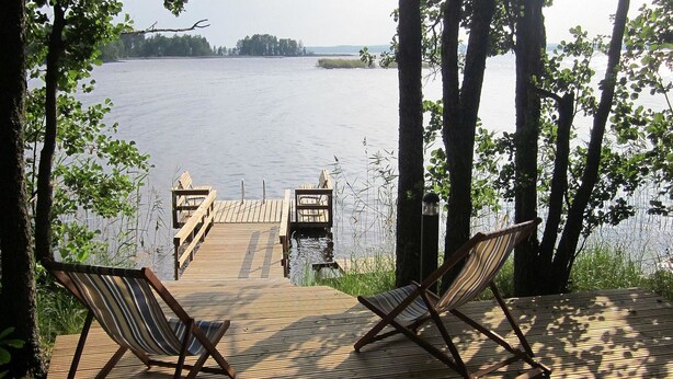 A wooden deck with two striped lounge chairs facing a calm lake, surrounded by trees. - Villa raita by Interhome (Rääkkylä)