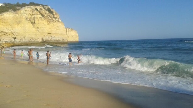Ein Strand mit Menschen, die am Ufer entlanggehen, Wellen, die auf den Sand treffen, und einer Klippe im Hintergrund.