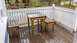 Een houten terras met een tafel en bankje, een witte reling, en een uitzicht op een chalet in de achtergrond.
