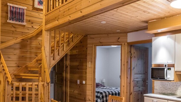 Een houten interieur met een trap, een eethoek met stoelen, een keuken met een magnetron, en een bed op de achtergrond.