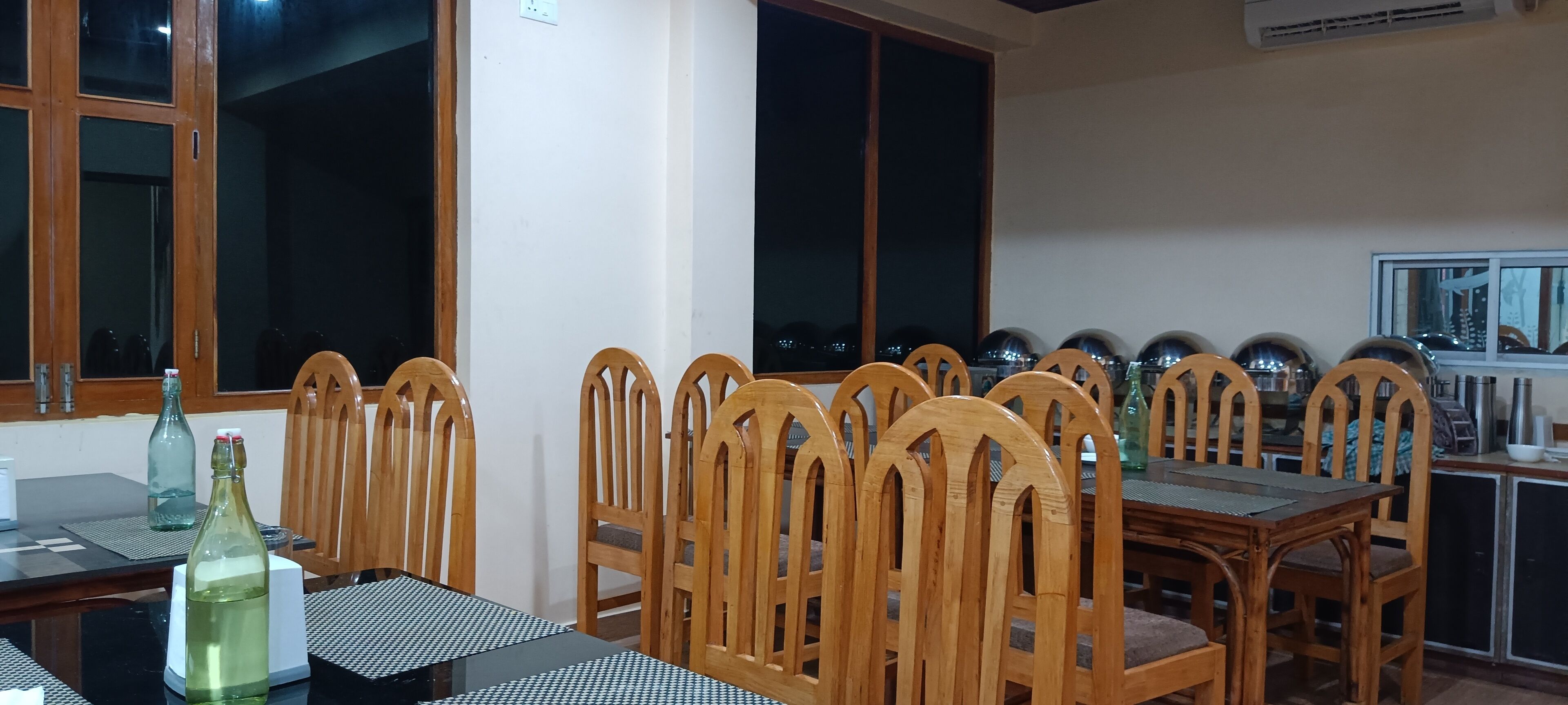 Un comedor con sillas de madera y una mesa, ventanas amplias, y una zona de cocina con armarios y electrodomésticos.