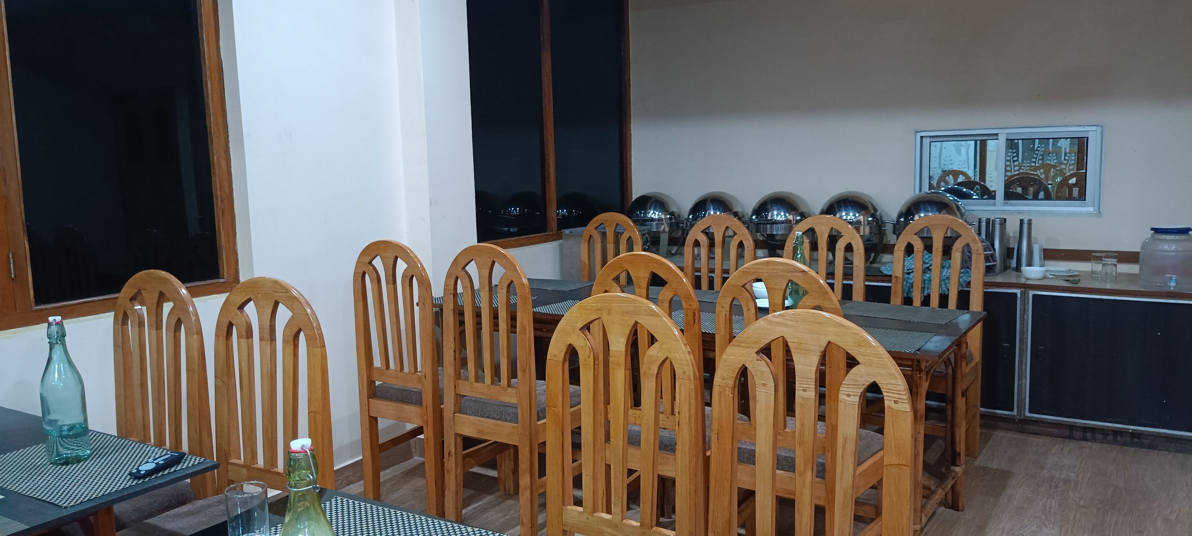 A dining area with wooden chairs and a table, large windows, and kitchen appliances in the background.