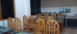 A dining area with wooden chairs and a table, large windows, and kitchen appliances in the background. - Blue Ocean Resort (Port Blair)