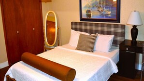 A hotel room with a bed, a wooden wardrobe, a mirror, a painting of boats, and a nightstand with a lamp.