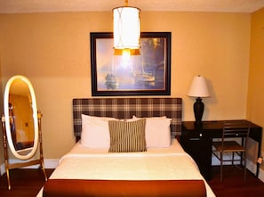 A hotel room with a bed, a desk, a chair, a mirror, and a painting on the wall.