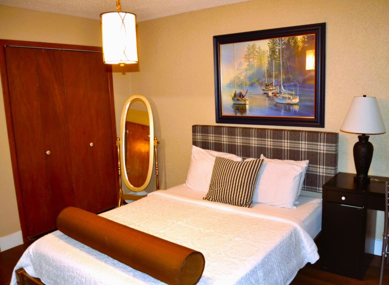 A hotel room with a bed, a wooden wardrobe, a mirror, a painting of boats, and a nightstand with a lamp.