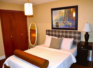 Habitación de hotel con cama, armario de madera, espejo, pintura de barcos y mesita de noche con lámpara.