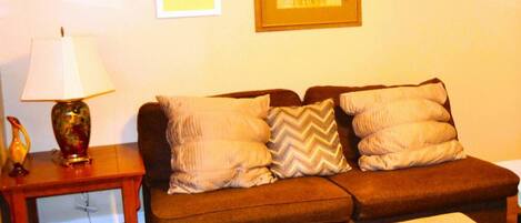 A cozy living room with a brown sofa, a matching ottoman, a wooden side table with a lamp, and two framed artworks on the wall.