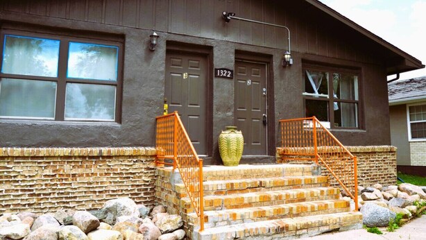 A two-story building with a stone foundation, a large green vase on the front steps, and a number "1322" on the door.