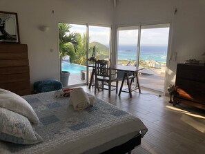 Un dormitorio con una cama grande, un aparador de madera, una silla y una mesa. Se ve una piscina y el océano a través del ventanal corredero.