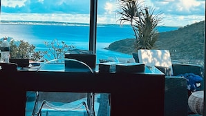 Een kamer met een groot raam met uitzicht op de kust, een bureau met stoel, en een potplant.