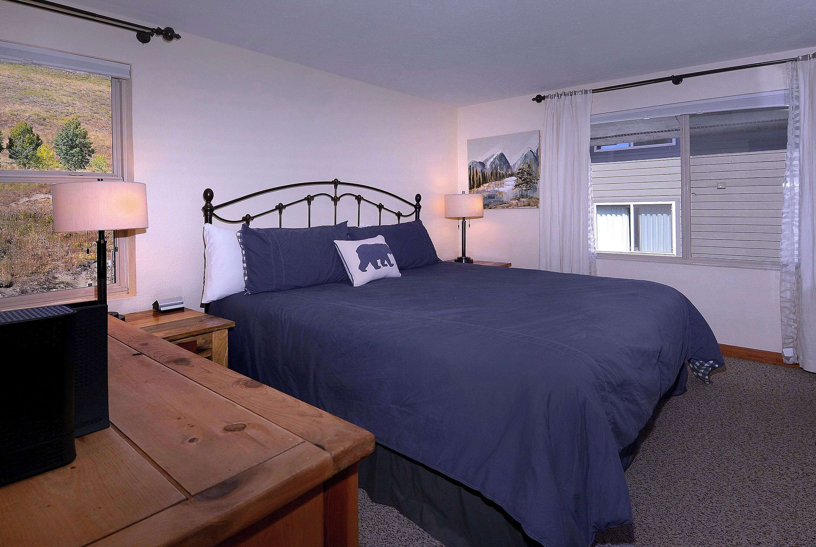 Une chambre à coucher avec un lit, deux tables de chevet, une commode en bois, une fenêtre avec des rideaux et un tableau au mur.