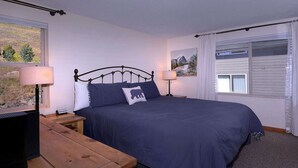 Quarto com uma cama, duas mesas de cabeceira, uma cômoda de madeira, uma janela com cortinas e um quadro na parede.