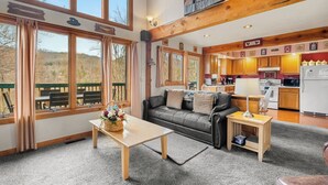 A spacious living area with a large window, a wooden table, a black leather sofa, and a kitchen in the background. - Summer Retreat - Treetop View Cabin with Hot tub & Pool Table (Gatlinburg)