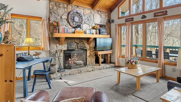 A cozy living room with a stone fireplace, wooden beams, a television, and a large window overlooking a deck. - Summer Retreat - Treetop View Cabin with Hot tub & Pool Table (Gatlinburg)