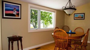 A dining room with a round wooden table and six chairs, a window with a view of trees, and two framed pictures on the walls. - Waterfront Vacation Home-4 bedrooms-2.5bath-Laundry room inside-2 heat pumps (York)