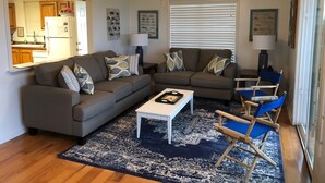 A living room with a gray sofa, a coffee table, and a blue armchair. There is a ceiling fan and a window with blinds. - Camp PJs - Coastal Realty Group (Grand Isle)