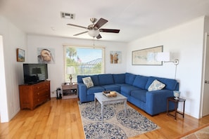 A living room with a blue sectional sofa, a wooden entertainment center, a ceiling fan, and a window with blinds.