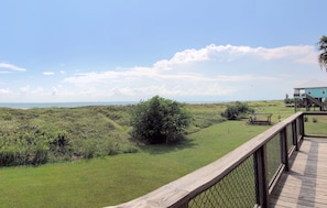 A wooden deck with a chain-link fence, a grassy area with bushes, and a distant view of a building with a staircase.