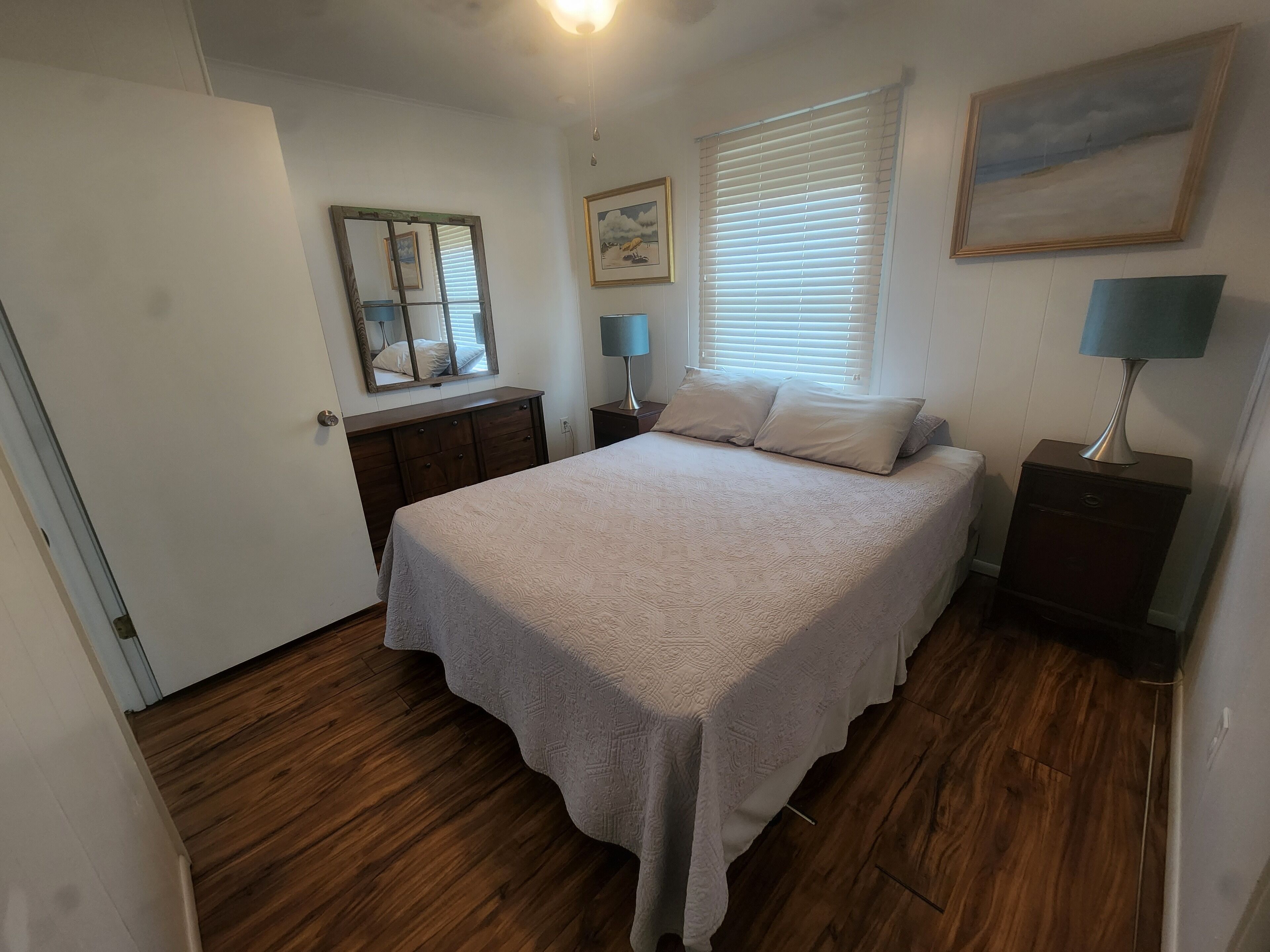 A bedroom with a bed, a dresser, a nightstand, a lamp, a window with blinds, and a painting on the wall.