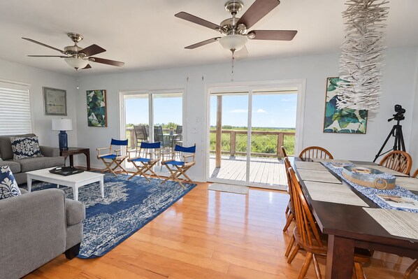 A spacious living room with a large wooden dining table, blue and white patterned rug, and ceiling fans. - Camp PJs - Coastal Realty Group- Beachfront (Grand Isle)