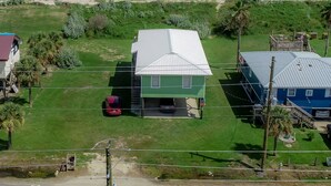 A house with a green roof and a garage, surrounded by a grassy yard and palm trees, with a beach and ocean in the background. - Camp PJs - Coastal Realty Group (Grand Isle)