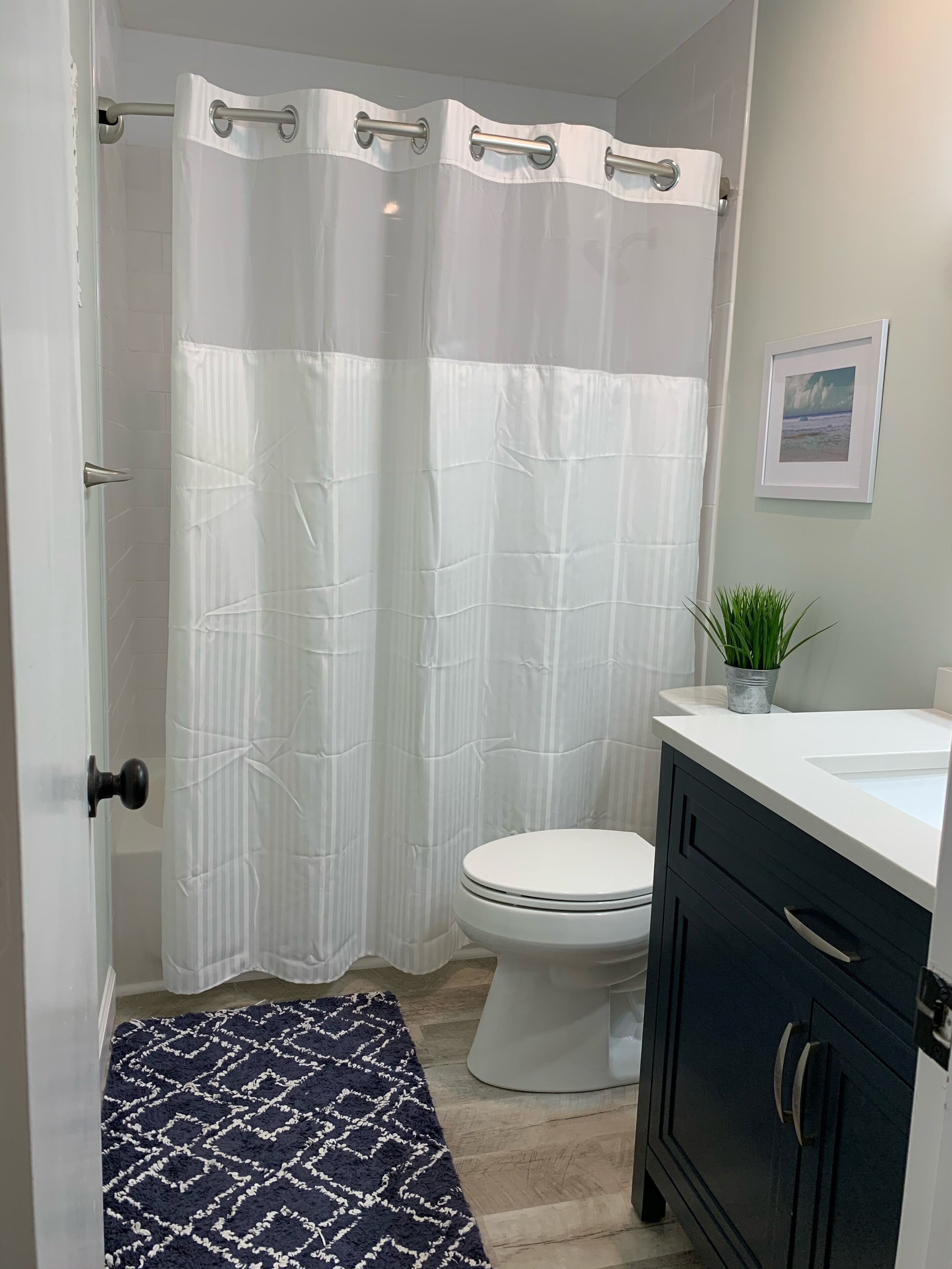 A bathroom with a white toilet, a white shower curtain, a black rug, and a small potted plant.