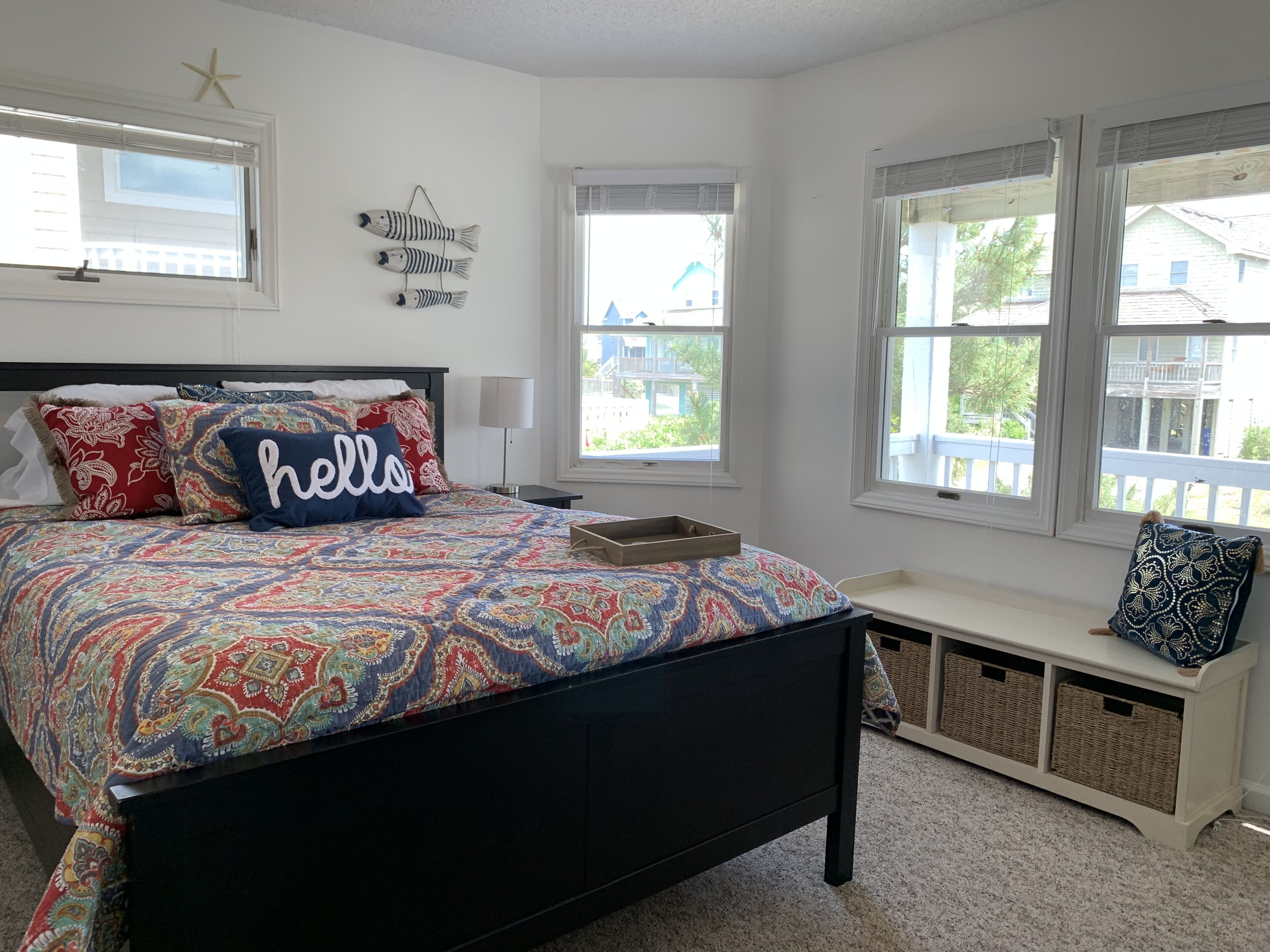 A bedroom with a patterned bedspread, a bench with baskets, and a view of a house through the windows.