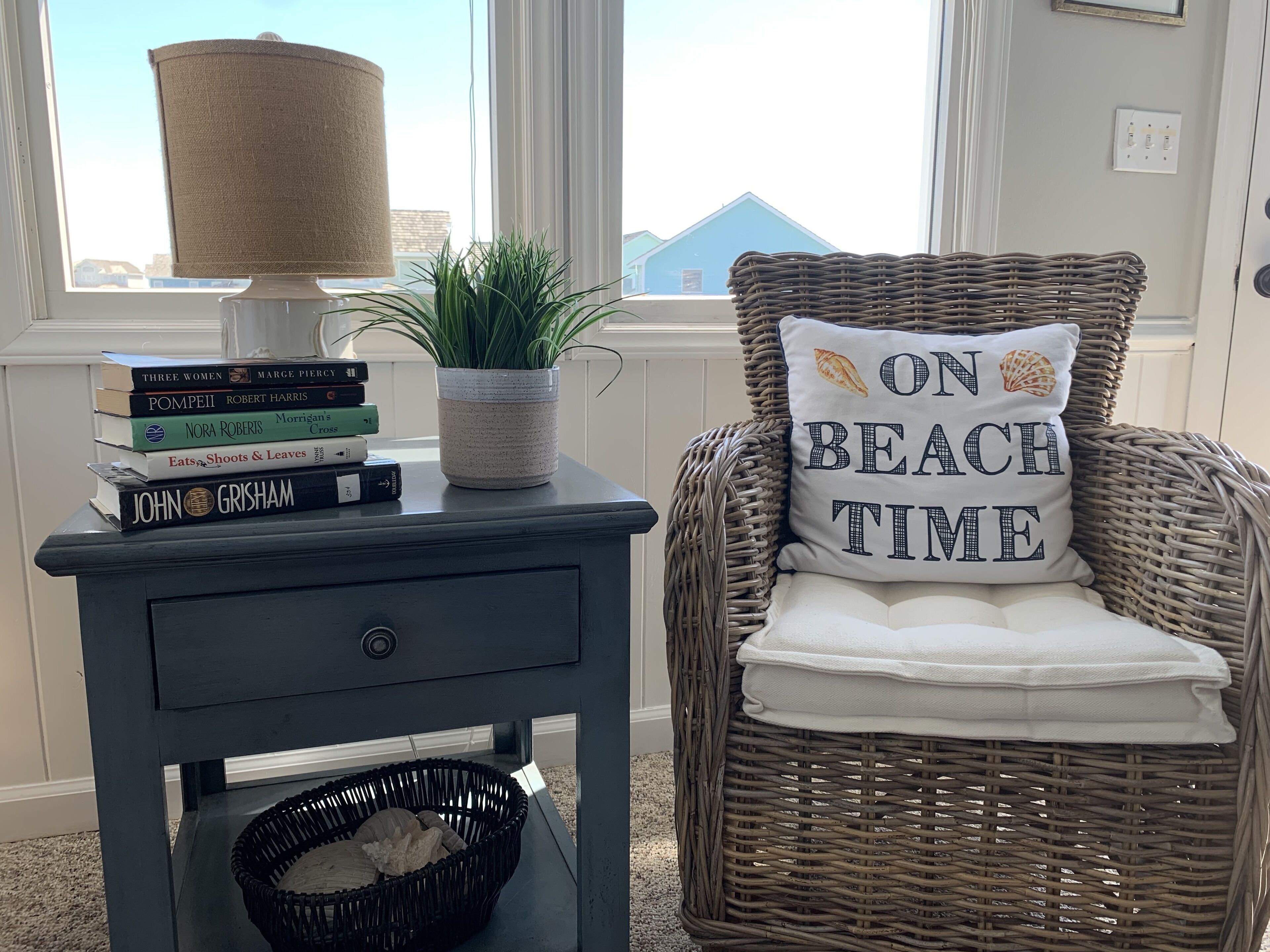 Una habitación con una silla de mimbre, una mesita con una lámpara, una planta y una pila de libros.
