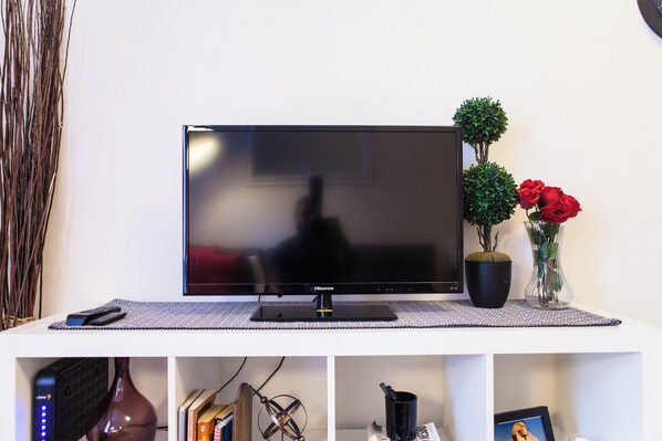 A flat-screen TV on a white stand with a remote control, a decorative vase with red roses, and a small potted plant. - Artist Modern 2 Bedroom Time Square / Central Park Apartment (Manhattan)