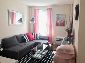 Un salón con un sofá gris, una mesita de centro pequeña, un tapete a rayas blanco y negro, una silla rosa y un jarrón con flores rojas.