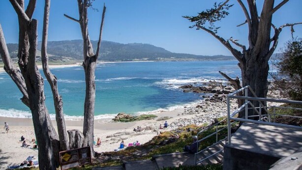 A beach with rocky terrain and trees, a staircase leading down to the beach, and a clear view of the ocean.