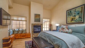 A spacious bedroom with a large bed, a fireplace, a bench with cushions, a TV, and a painting on the wall.