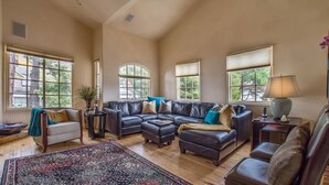 A spacious living room with a sectional sofa, a small armchair, and a coffee table. There are large windows providing natural light and views of the outdoors.